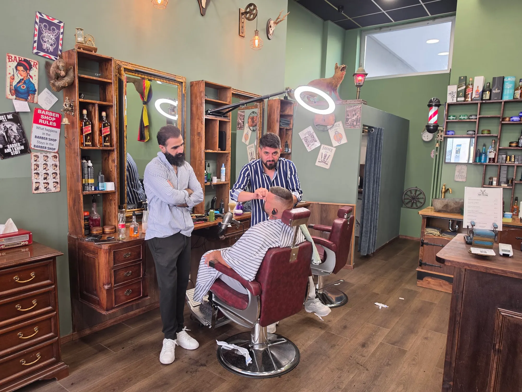 Zwei Barber bei der Arbeit an einem Kunden im roten Barberstuhl, Holzregale und Spiegel im Hintergrund